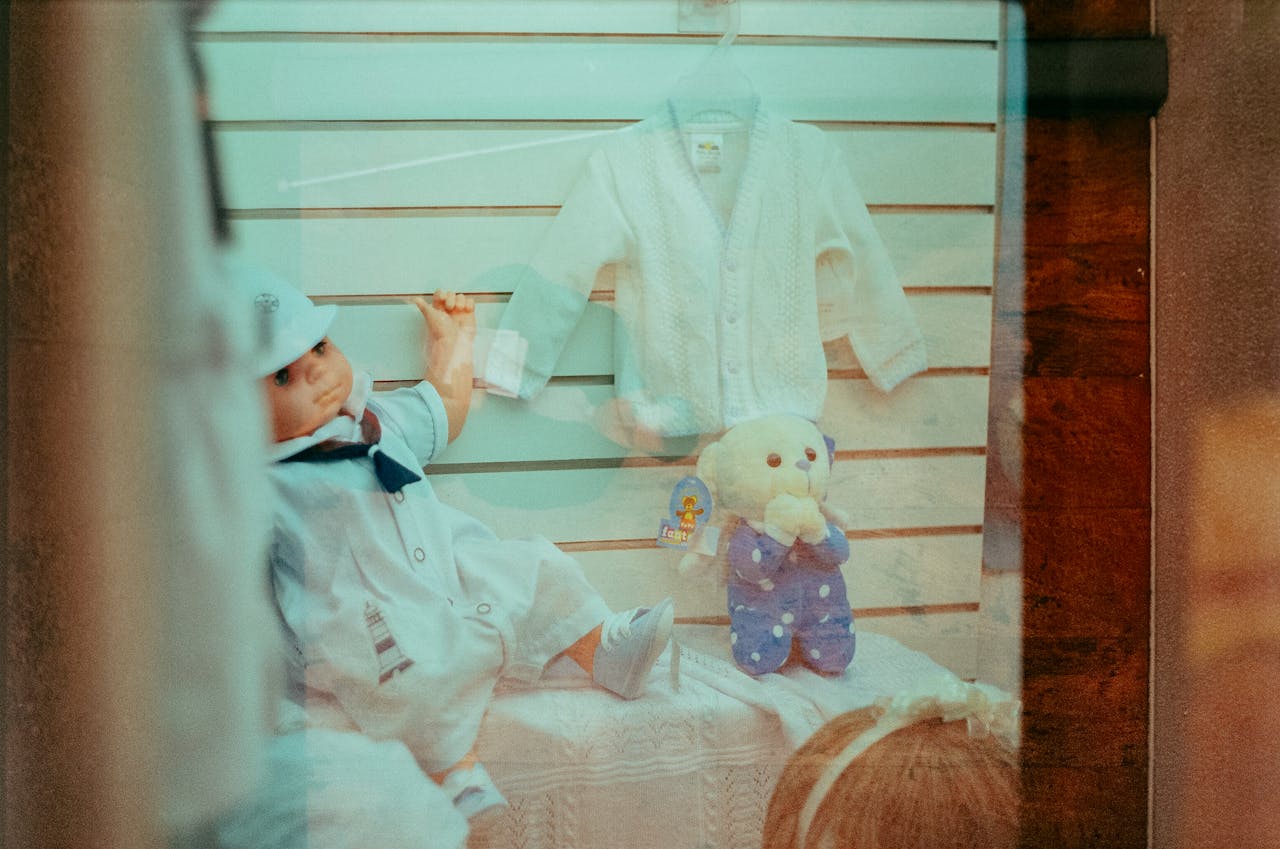 Storefront display featuring a cute doll, teddy bear, and baby clothes in soft colors.