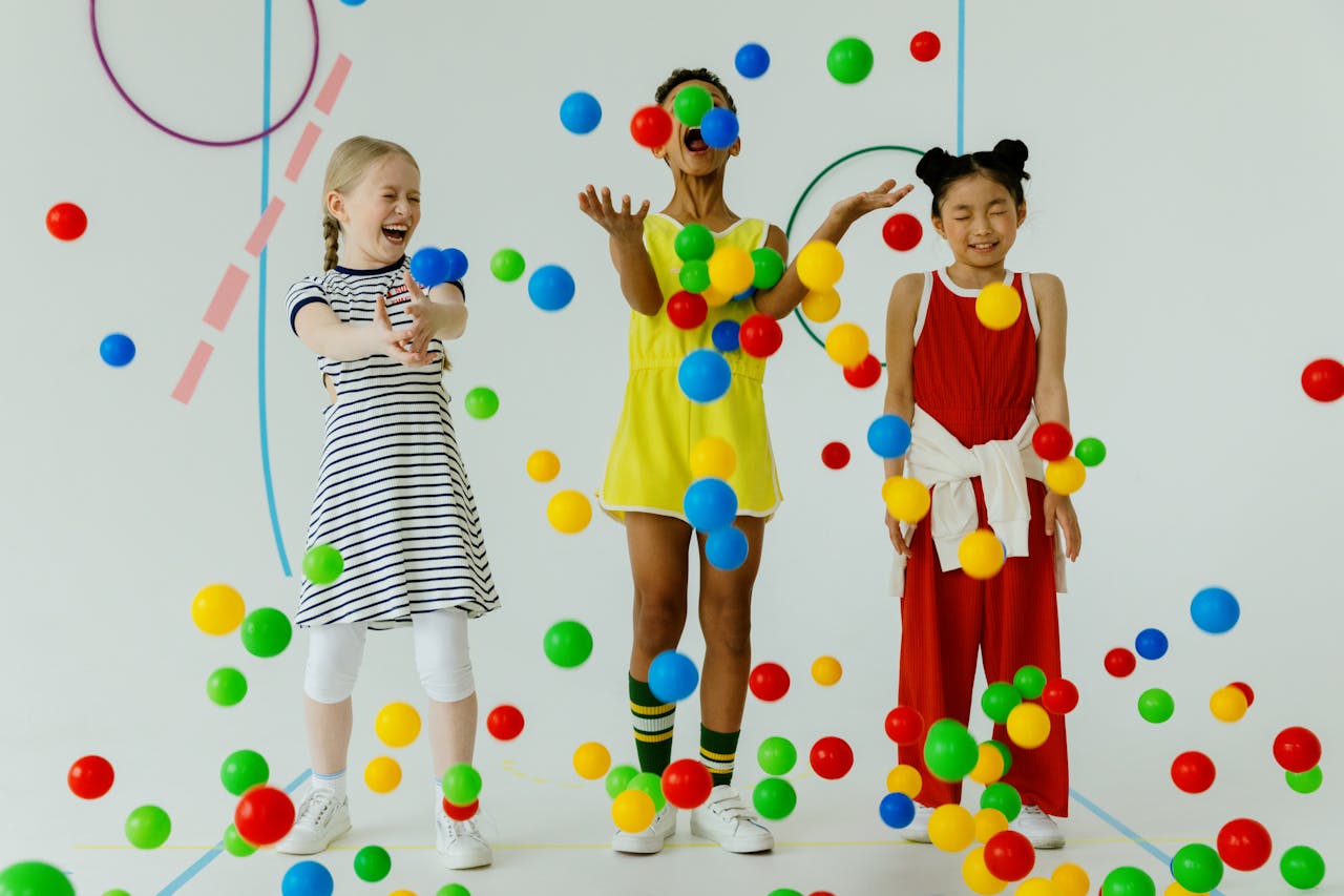 Three diverse girls laughing and playing with colorful balls indoors.